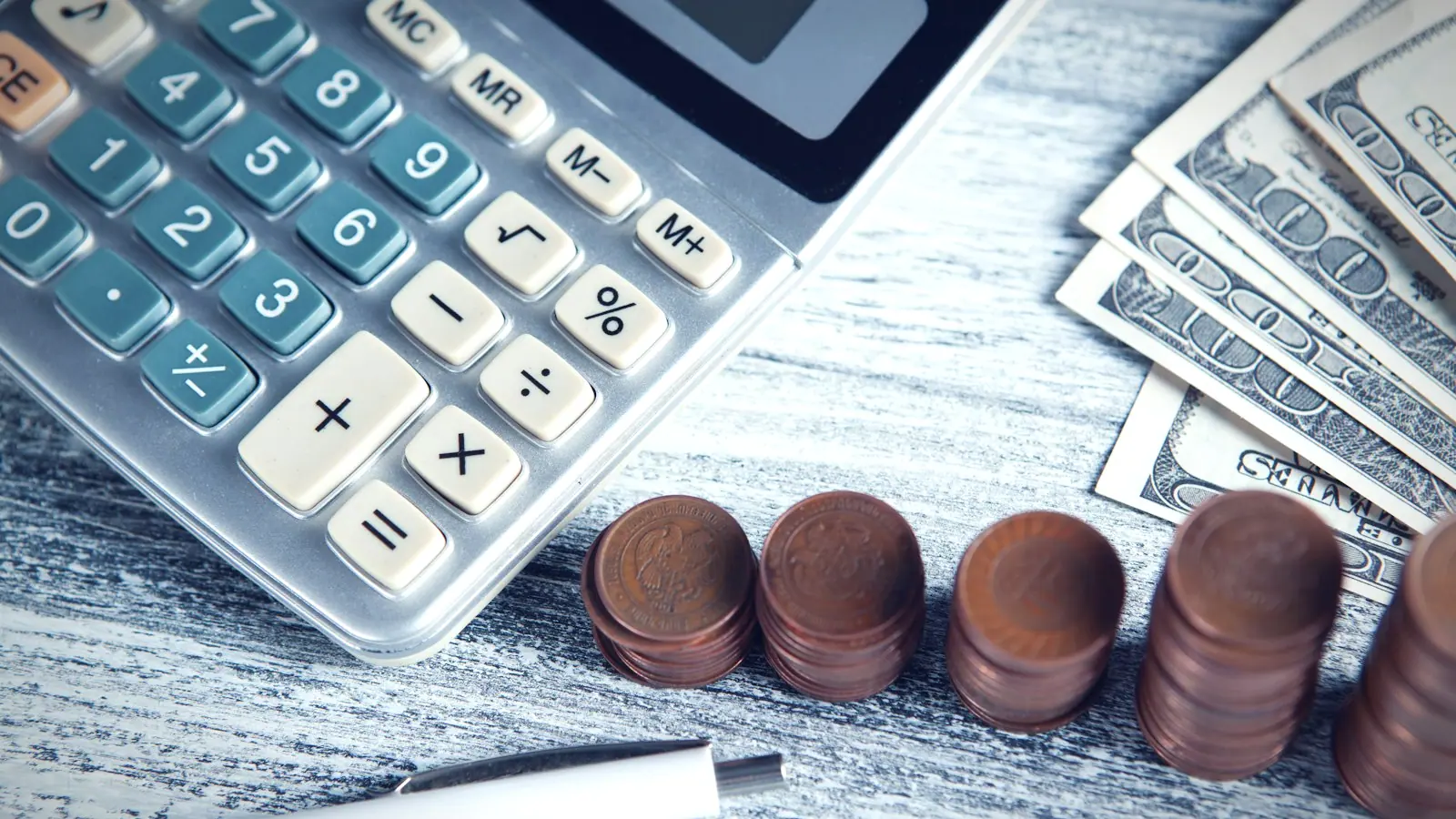 A calculator and pen beside stacked coins and banknotes on a desk.