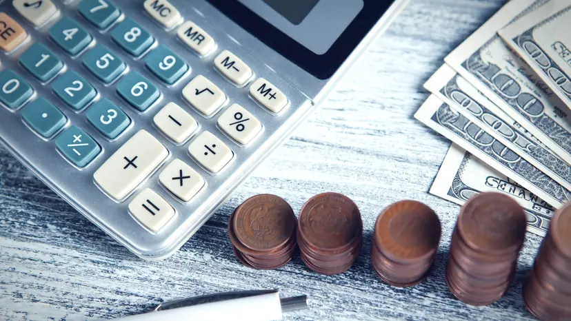 A calculator and pen beside stacked coins and banknotes on a desk.