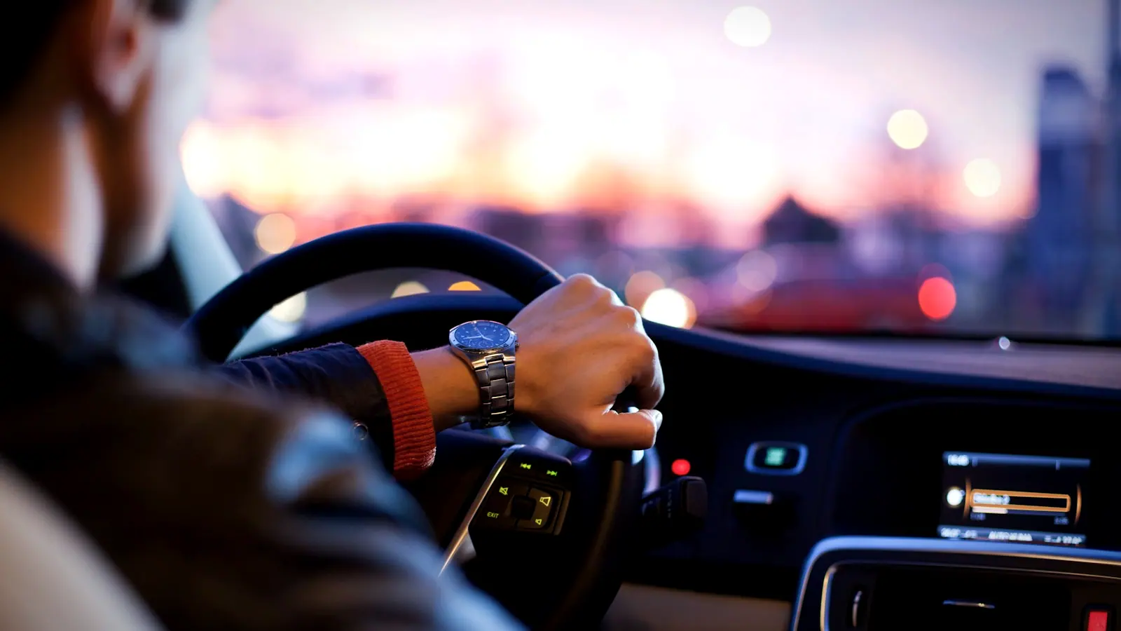 A driver's hand on the steering wheel at dusk with city lights blurred ahead.