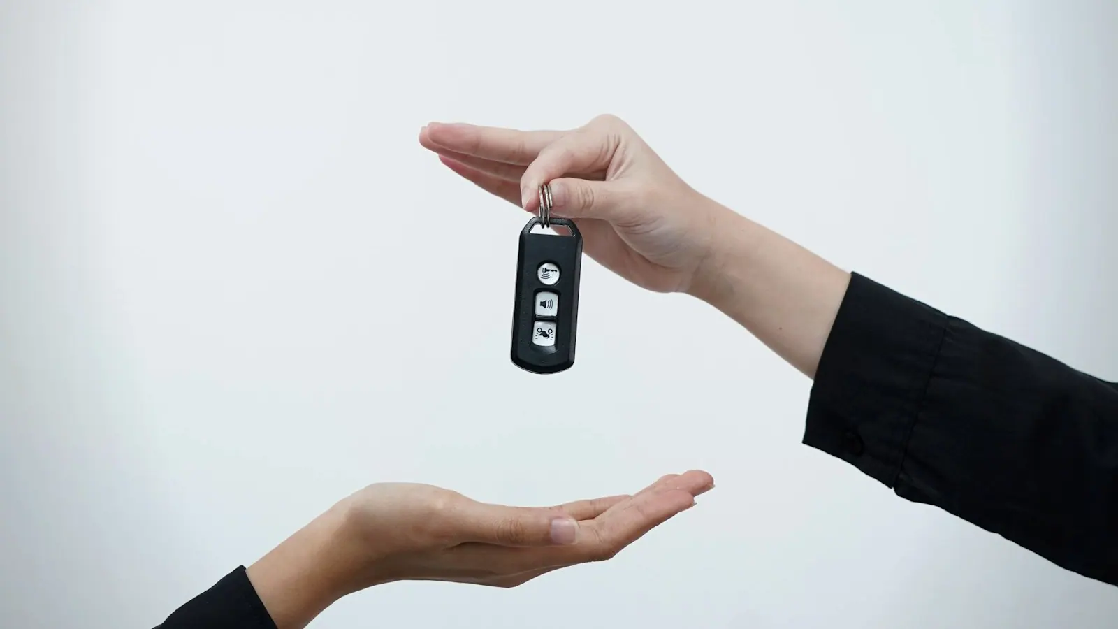 One hand passing a car key to another hand against a plain background.